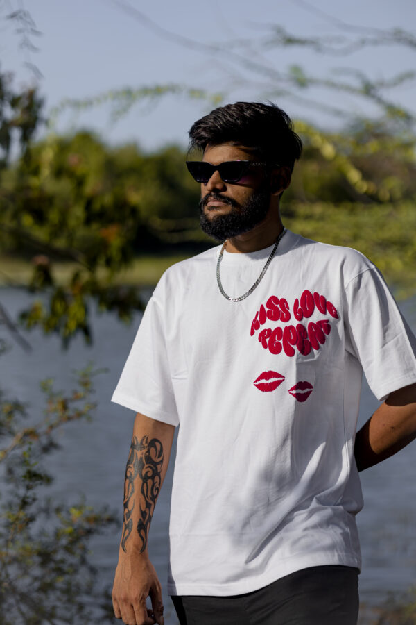 Man wearing a white oversized t-shirt with bold red text that reads "Kiss Love Repeat" and red lip print graphics, styled with sunglasses and a chain, standing by a lake.