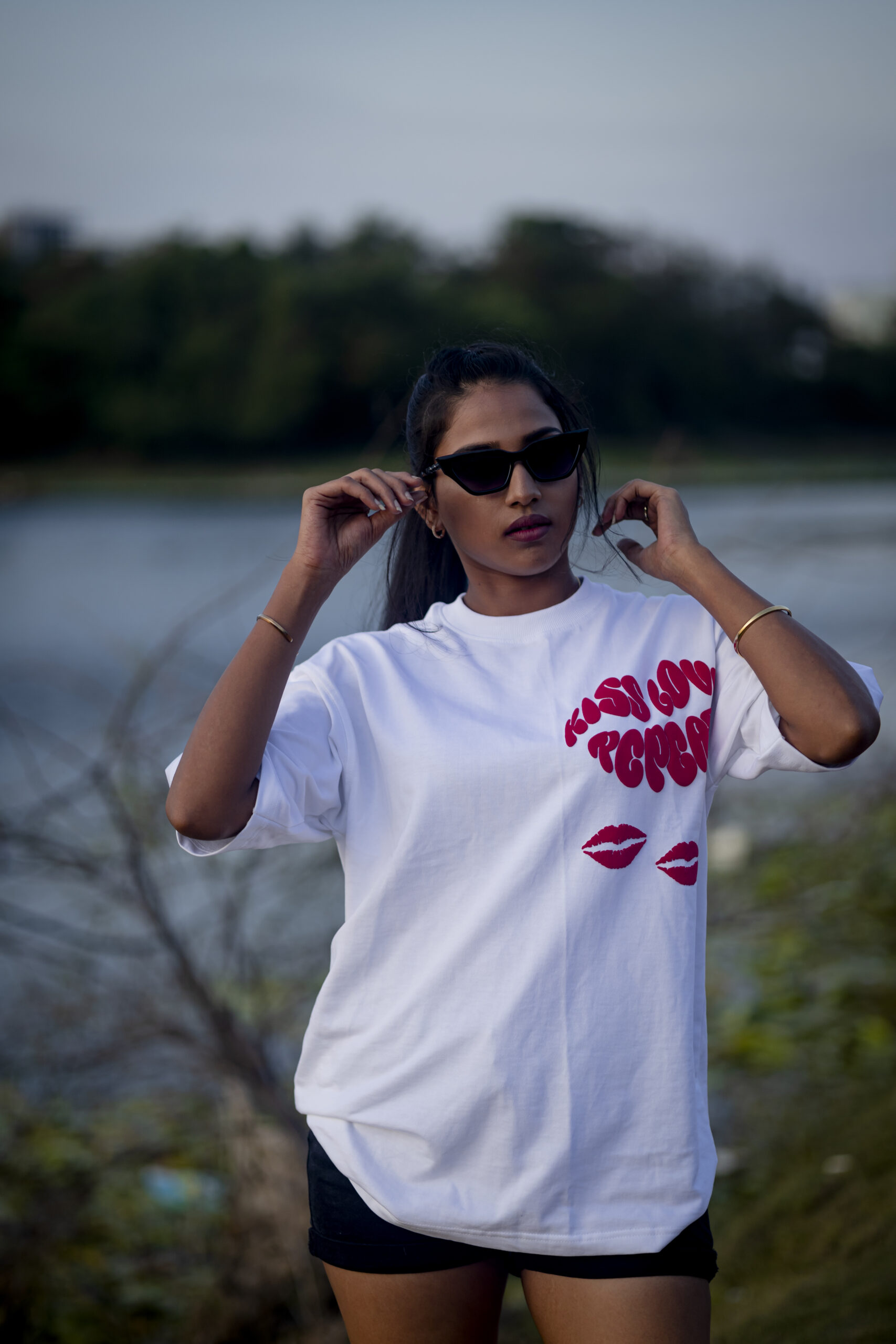 Woman wearing a white oversized t-shirt with bold red "Kiss Love Repeat" text and lip prints, adjusting her sunglasses by a lakeside.