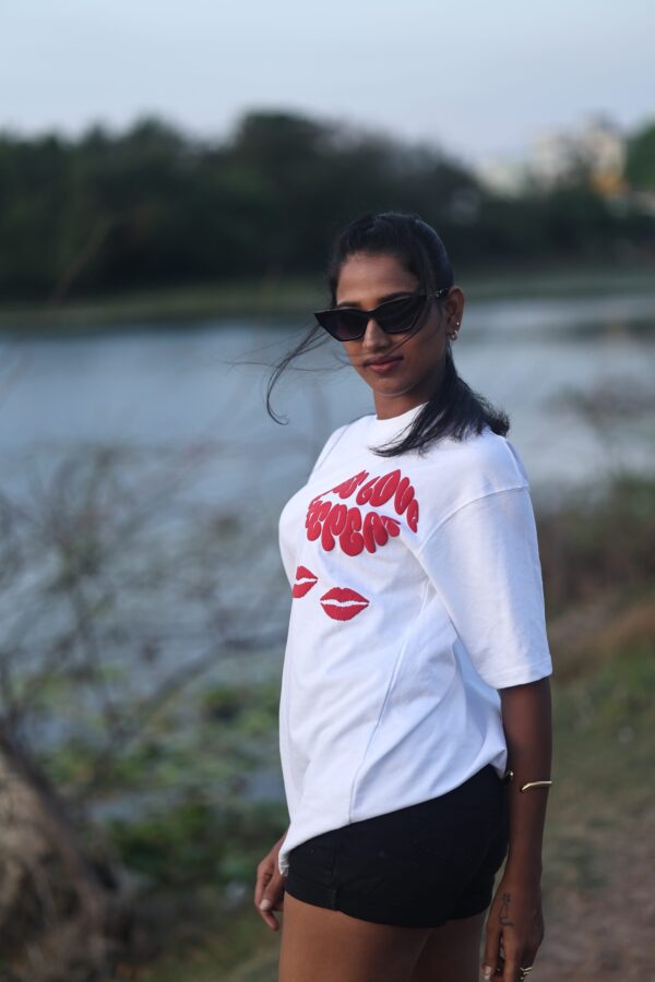 Confident woman in black shorts and white oversized t-shirt with red "Kiss Love Repeat" print, standing by the lakeside with wind-blown hair.