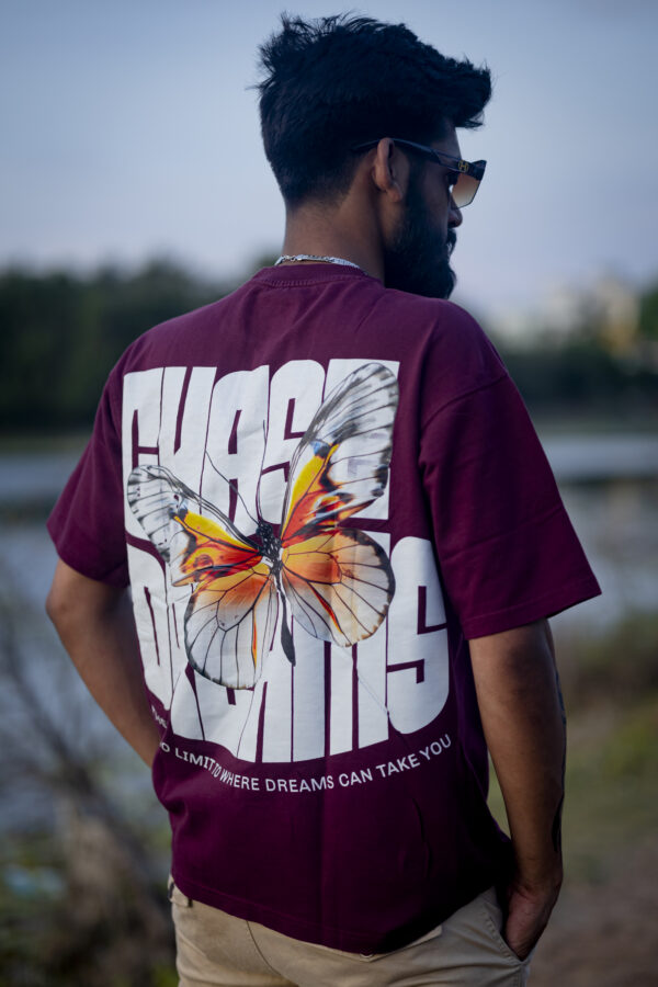 Man wearing a maroon oversized t-shirt with a large butterfly graphic and bold "CHASE DREAMS" text on the back, standing near a lake during sunset.