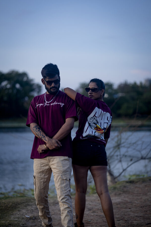 Man and woman in matching maroon oversized t-shirts posing near a lake; the man’s shirt reads "Escape the ordinary" and the woman's shirt features a bold butterfly graphic with "CHASE DREAMS" text.