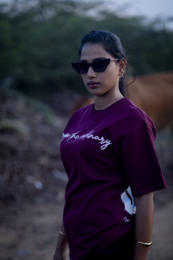 Woman wearing a maroon oversized t-shirt with the phrase "Escape the ordinary" printed on the front, styled with black sunglasses in an outdoor setting.
