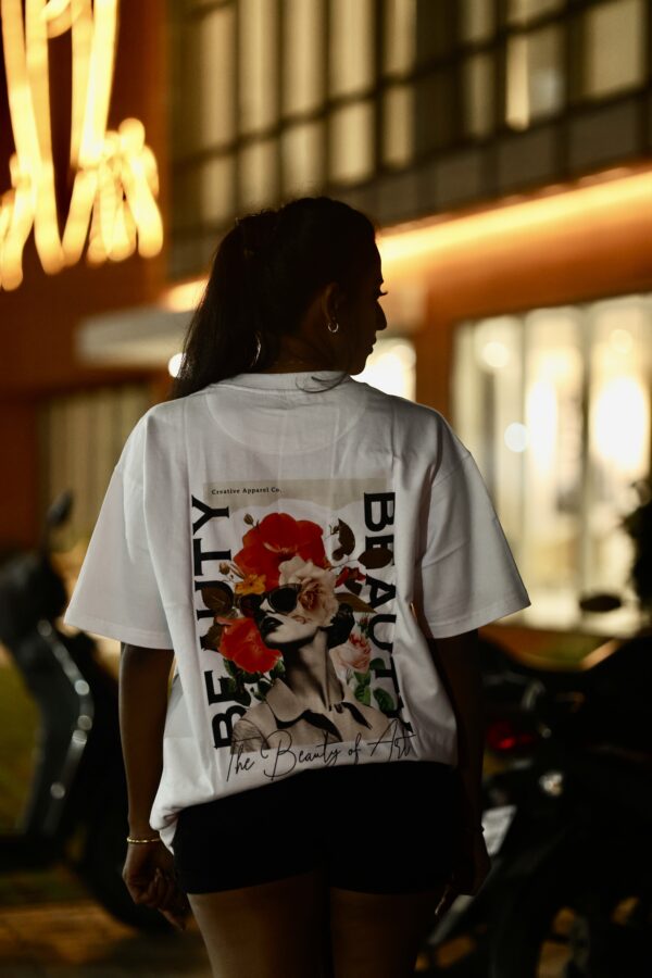A woman wearing a white oversized graphic t-shirt featuring a bold floral and portrait collage with the words “The Beauty of Art,” photographed at night in an urban setting.