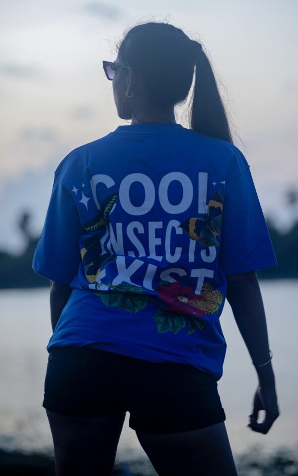 Woman facing the sunset near water, wearing a blue oversized Kläder t-shirt with bold “COOL INSECTS EXIST” text and colorful butterfly and flower graphics on the back.