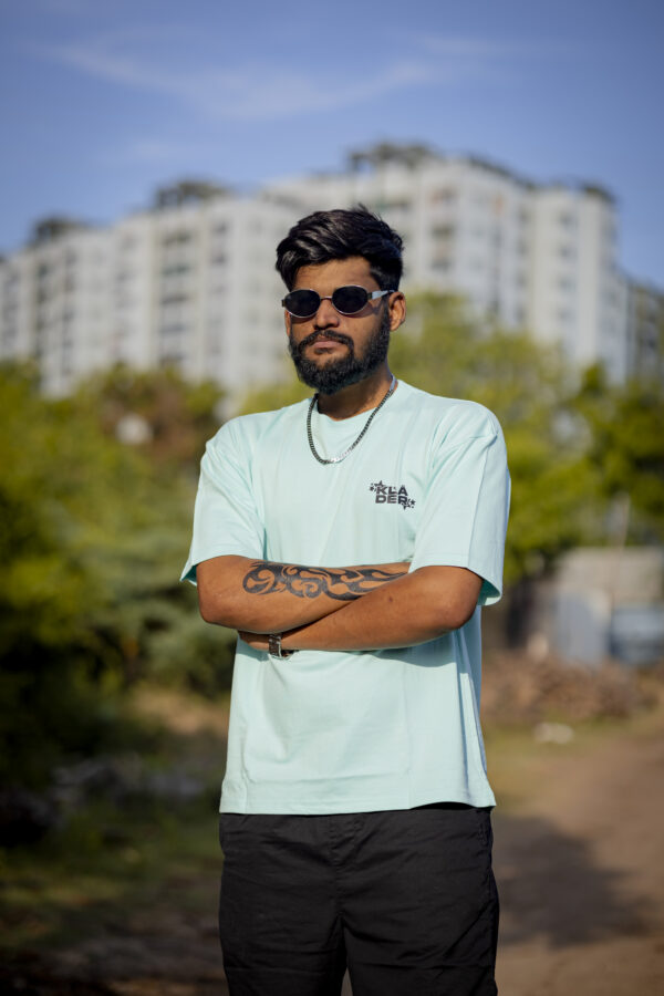 Man wearing a light blue oversized T-shirt by Kläder featuring a cute cartoon dinosaur dressed as a warrior knight on the back, styled with black shorts and accessories, outdoors with natural background.
