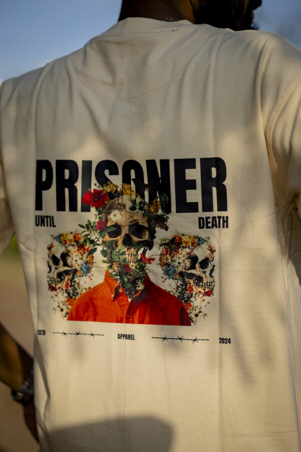 Back view of a beige oversized t-shirt featuring a skull and floral graphic with the text ‘PRISONER UNTIL DEATH,’ worn by a male model outdoors.