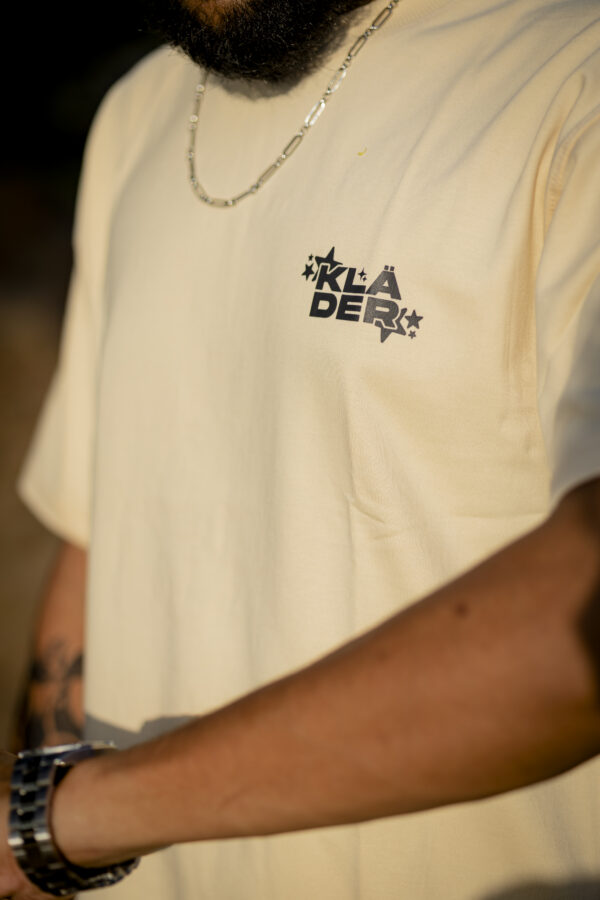 Close-up of the front of a beige oversized t-shirt with Kläder streetwear logo printed on the left chest, worn by a bearded male model