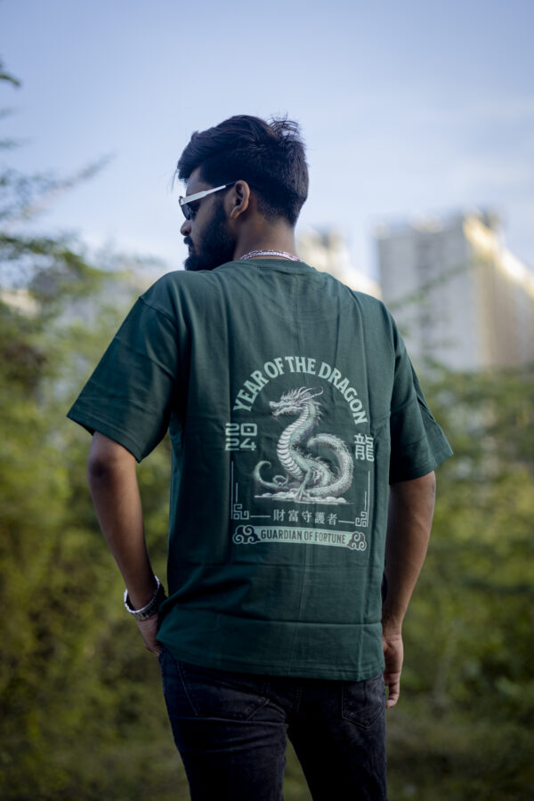 Man wearing a dark green oversized Kläder T-shirt featuring a bold dragon graphic with “Year of the Dragon 2024” and “Guardian of Fortune” text, standing outdoors with a cityscape in the background