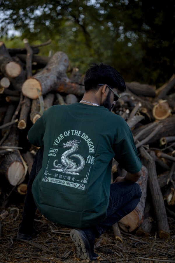 Man squatting in front of chopped logs wearing a forest green oversized Kläder T-shirt with a dragon graphic and “Year of the Dragon 2024 - Guardian of Fortune” text
