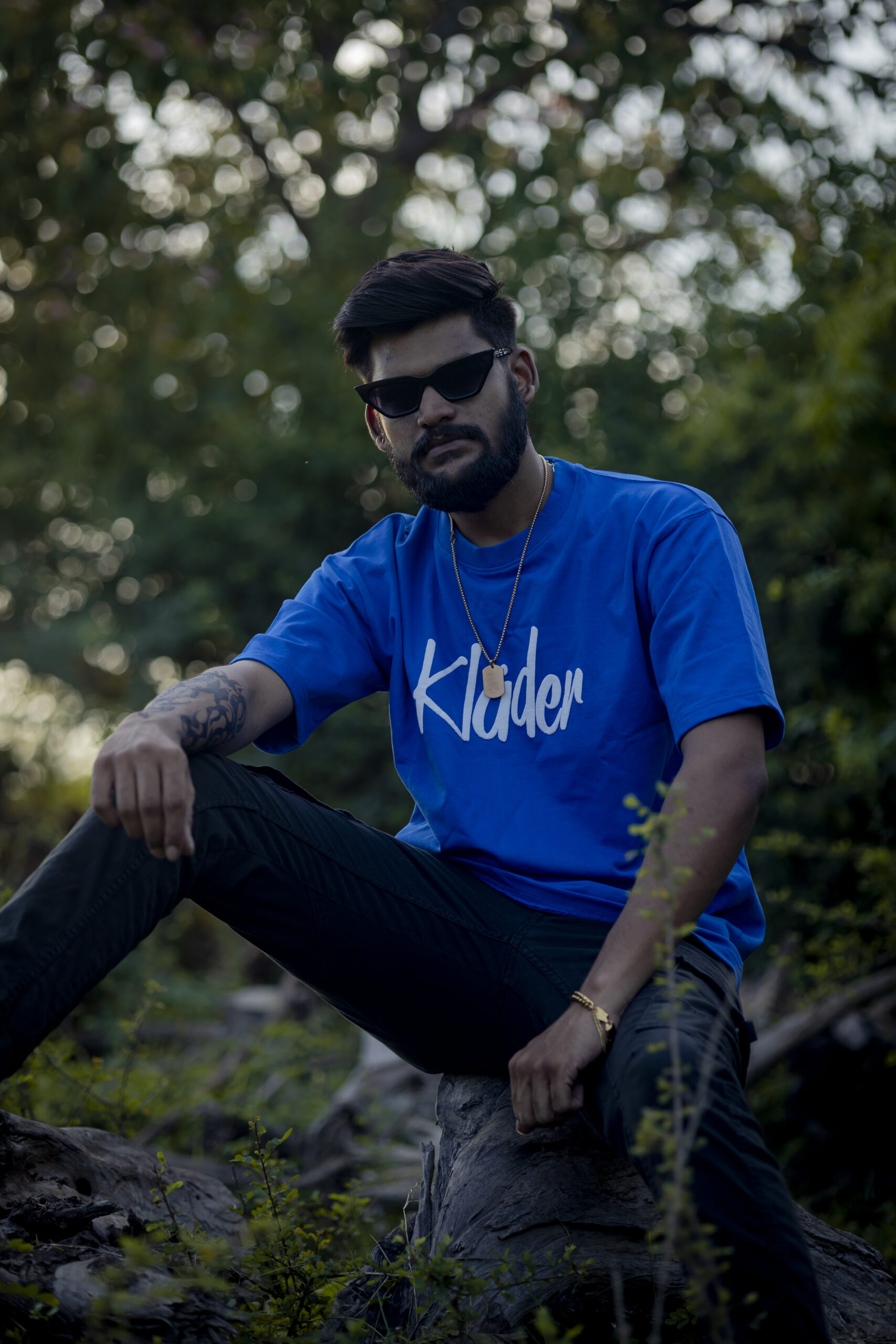 Man wearing a bold blue oversized Kläder t-shirt with white logo text, seated on a tree trunk in a forested outdoor setting, accessorized with sunglasses and a gold necklace.