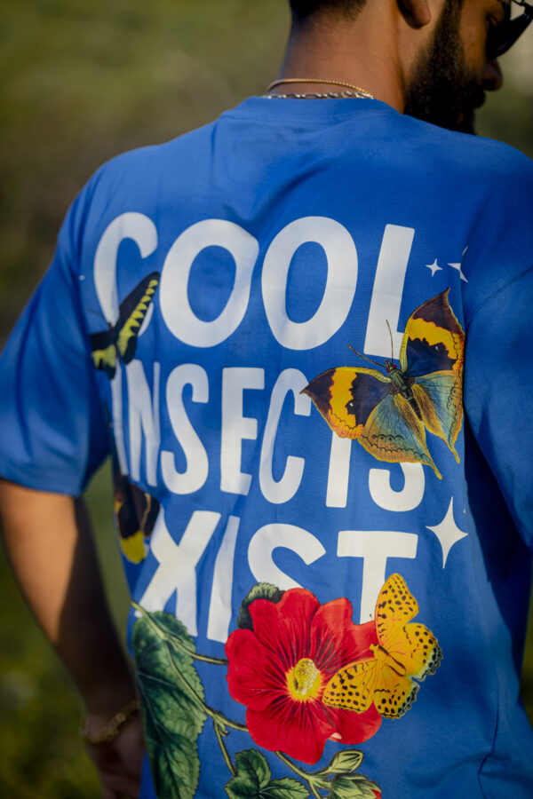 Back view of a person wearing a vibrant blue oversized unisex t-shirt by Kläder, featuring bold white text that reads “COOL INSECTS EXIST” with colorful butterfly and floral graphics.