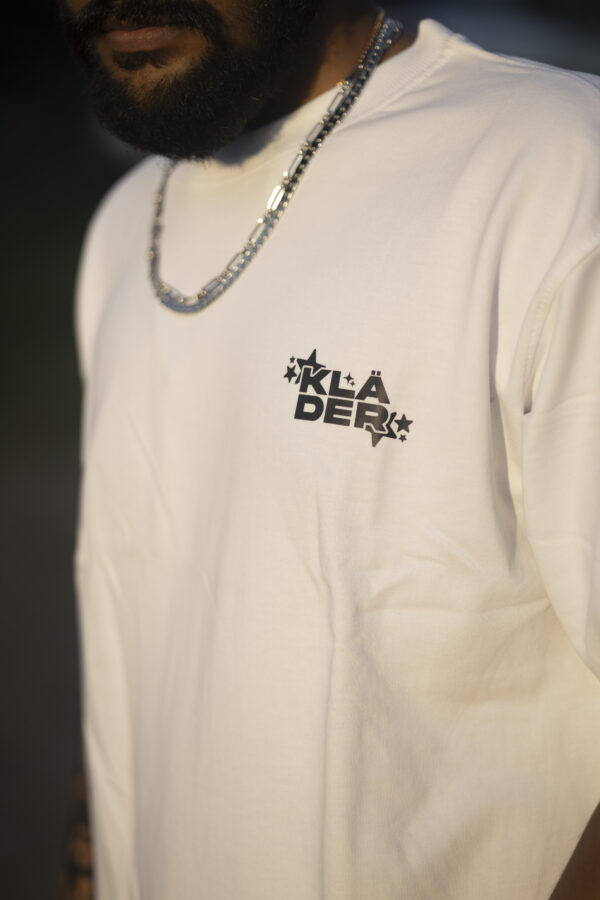 Man wearing a white T-shirt with a small 'KLÄDER' logo on the chest, standing outdoors with sunglasses and accessories
