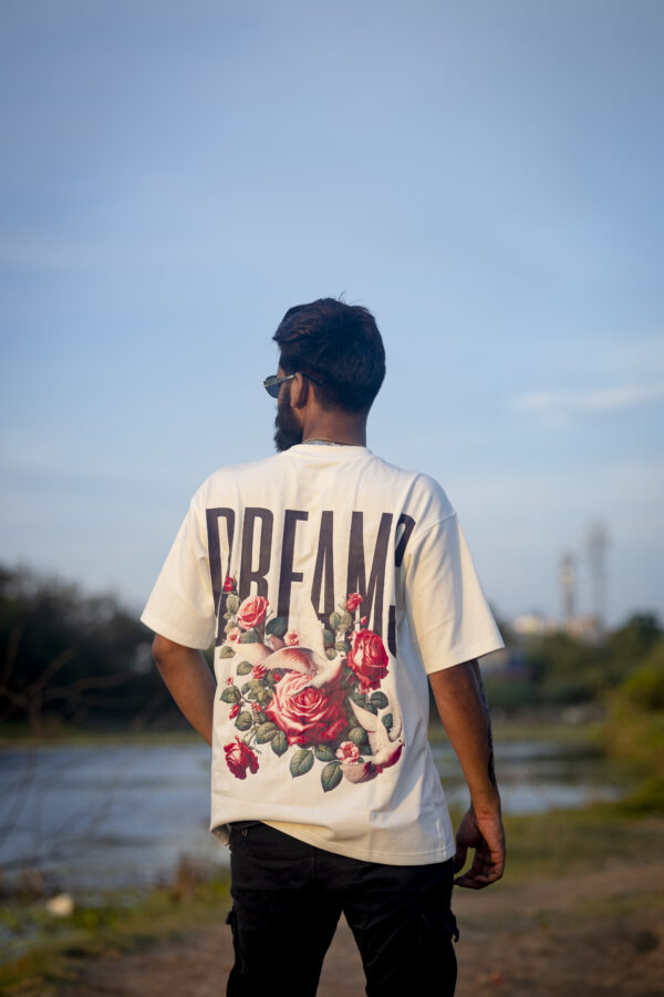 A man stands outdoors facing a serene riverside scene, wearing a white T-shirt with a bold back print featuring the word "DREAMS" and a vibrant floral and dove design.