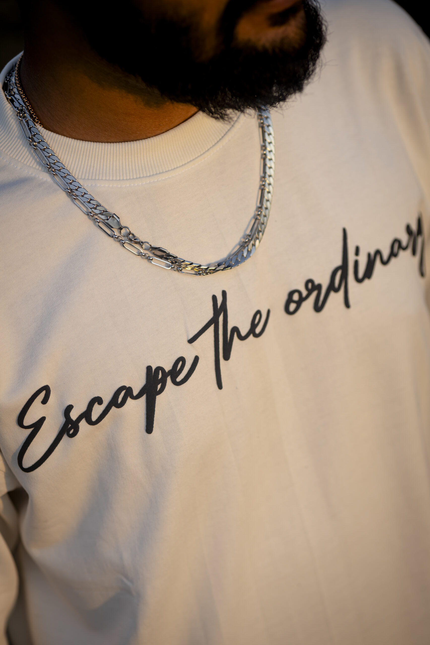Close-up of a man wearing a white T-shirt with the text "Escape the ordinary" printed in a handwritten style, paired with layered silver chains.