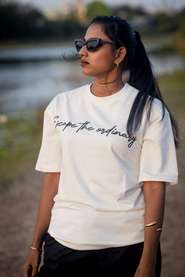 Woman wearing a white "Escape the ordinary" T-shirt, black sunglasses, and gold accessories, standing outdoors near a riverbank.
