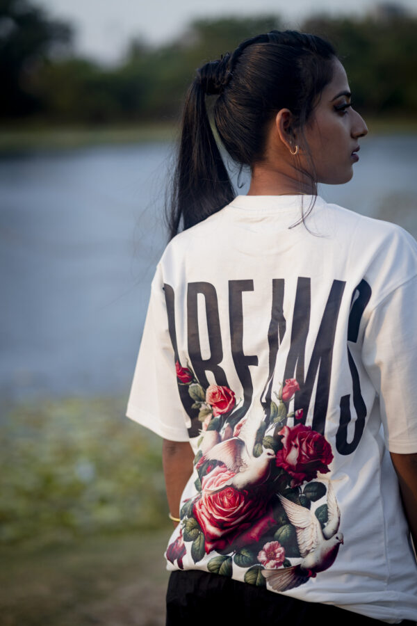 Woman wearing a white "DREAMS" graphic T-shirt with floral and dove design on the back, standing near a serene lakeside.