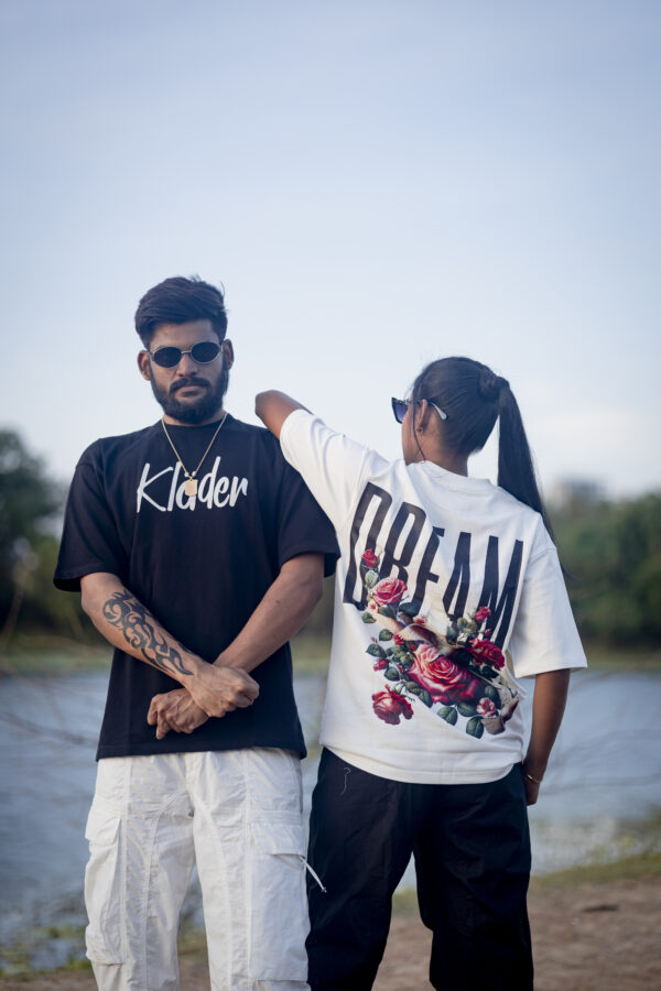 Man and woman wearing statement graphic T-shirts, with the man in a black klader tee and the woman in a white “DREAMS” tee with floral and dove print on the back, posing outdoors near water.