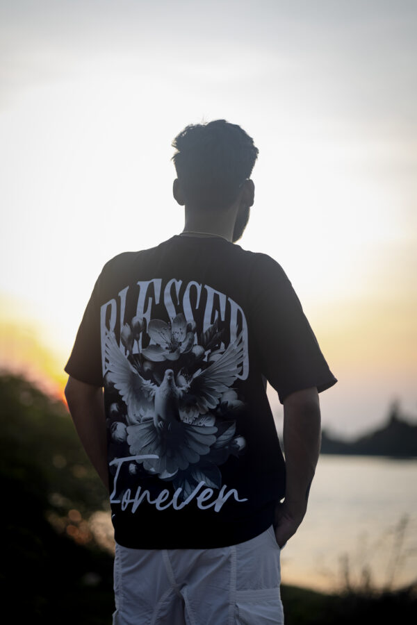 Man facing sunset, wearing black oversized t-shirt with dove and flower graphic and "Blessed Forever" text.
