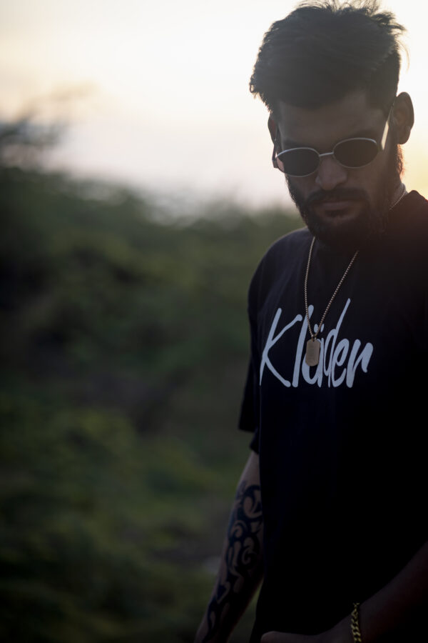 Bearded man wearing sunglasses and a black klader graphic t-shirt at sunset, exuding a bold streetwear look.