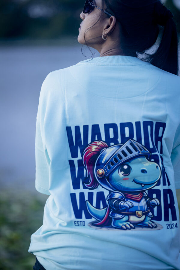 Woman wearing a light blue oversized Kläder T-shirt featuring a cartoon dinosaur warrior print on the back, styled casually with black shorts and sunglasses, posing outdoors in natural light
